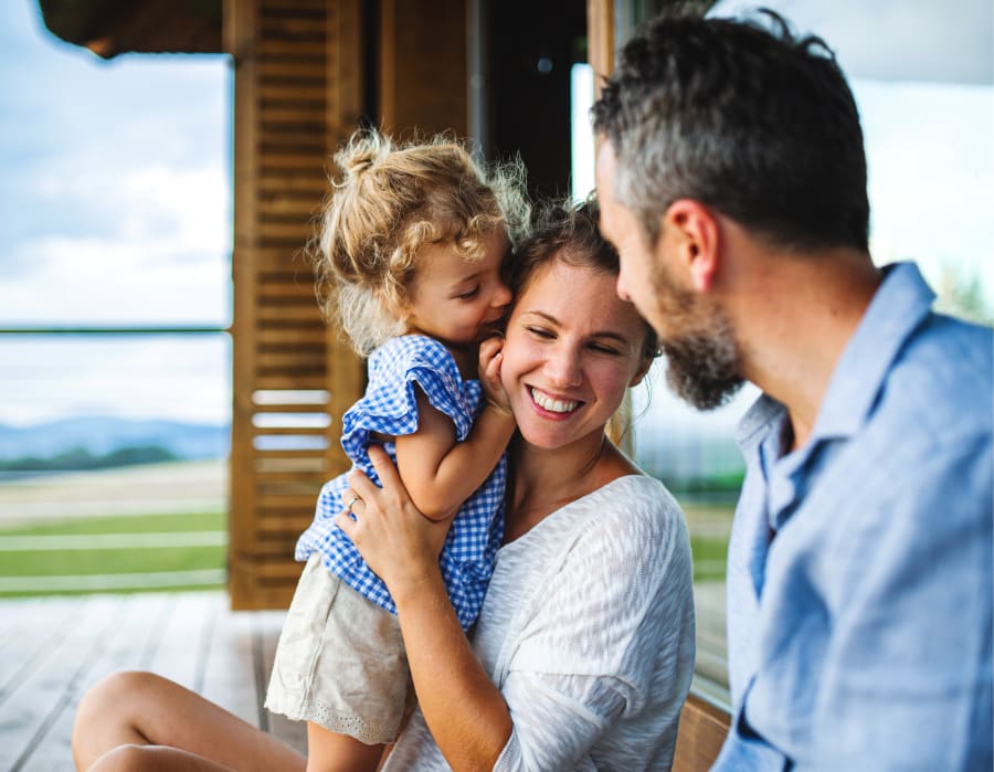 Family sitting outside on a porch. Woman smiling with child standing in lap with grass and water in the background.